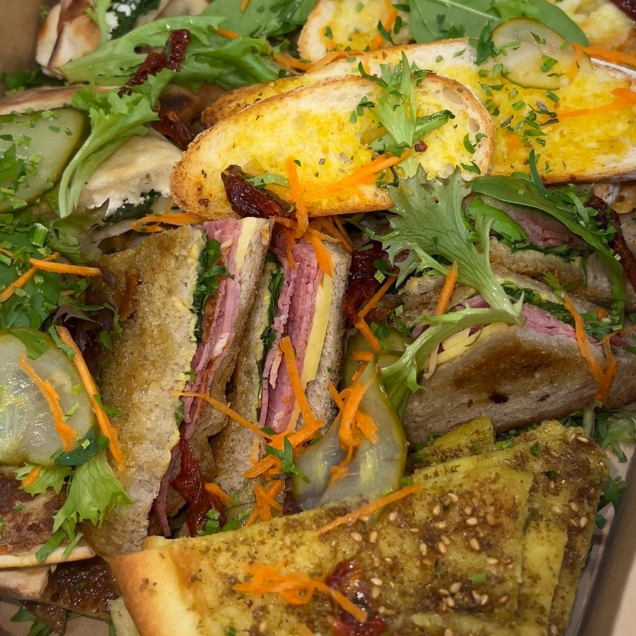 Close-up of a catering platter with garlic bread, sandwich quarters filled with sliced meat and cheese, and za'atar flatbread in a kraft box